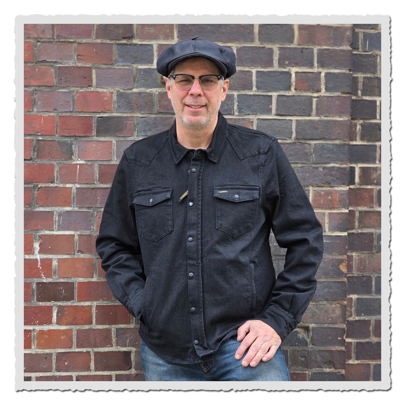 erson von vorn in dem schwarzen Rider Shirt-Hemd und dunkler Mütze vor roter Backsteinwand, eine Hand in der Tasche.